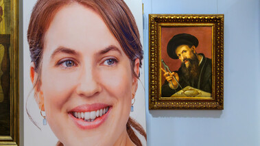 Frau vor klassischem Porträtgemälde, beide Gesichter in Blickrichtung Kamera.